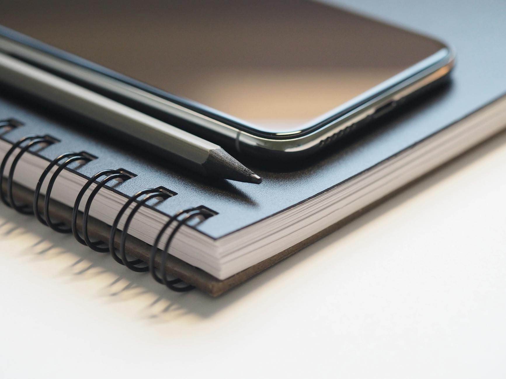 closeup photo of black smartphone near black and grey pencil on black spiral notebook