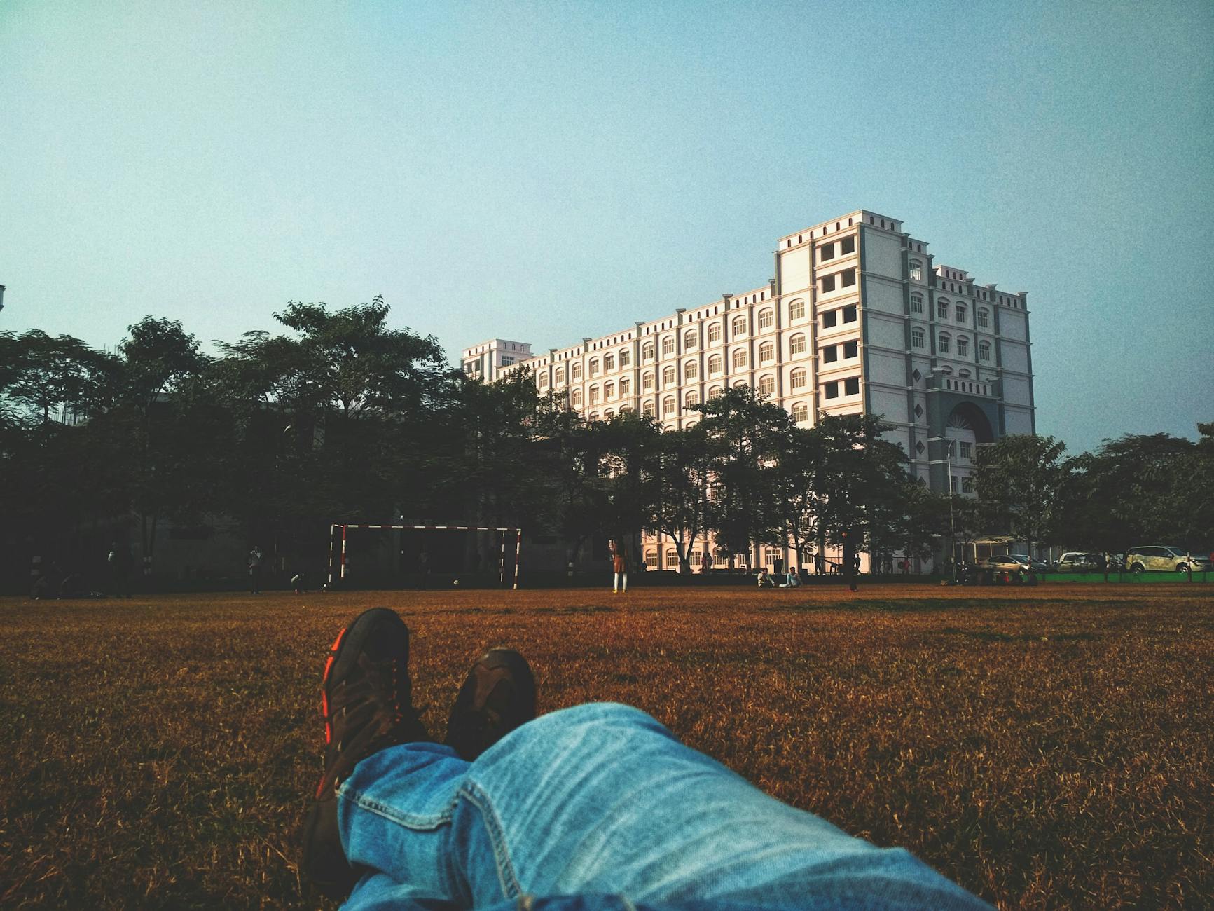 person in blue denim jean lying on brown grass field looking at white multi storey building